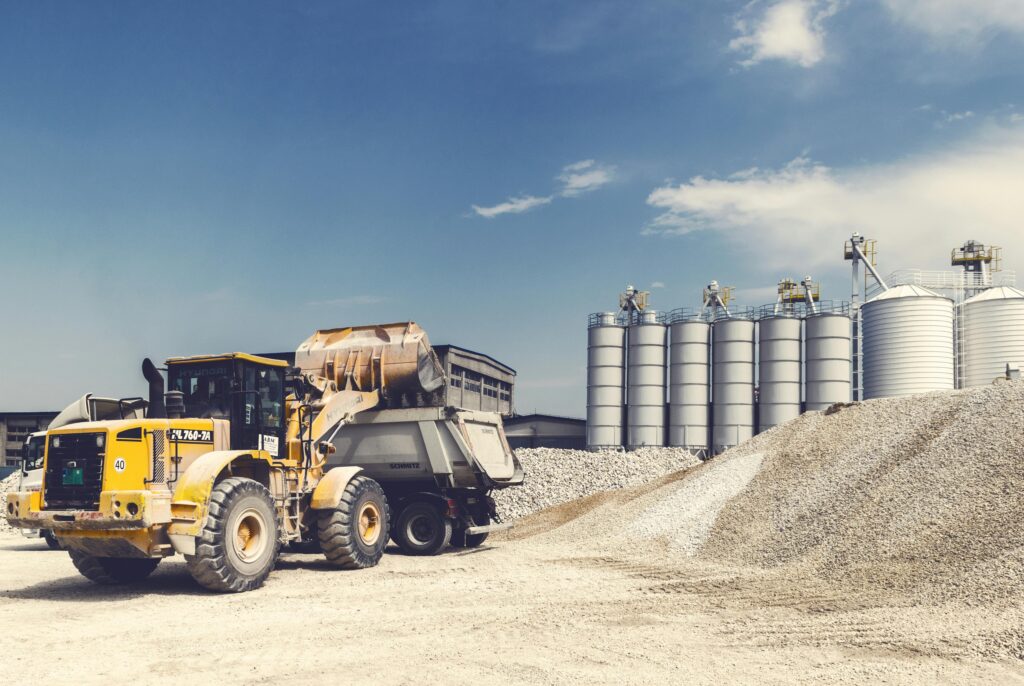 Yellow loader and truck operating at a gravel yard with silos in the background on a sunny day.